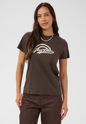 Kobieta z długimi brązowymi włosami ma na sobie brązowy t-shirt z grafiką "Good Vibes", brązowe spodnie i srebrny naszyjnik, stojąc na jednolitym tle.