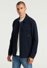 Marineblaues Jacket mit vier Fronttaschen, einem knöpfbaren Design und einem weichen, strukturierten Stoff, getragen über einem weißen Hemd und kombiniert mit blauen Jeans.