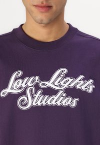 Ljubičasti dukserica s okruglim izrezom i izvezenim natpisom "Low Lights Studios" u bijeloj boji, koja prikazuje glatku teksturu.