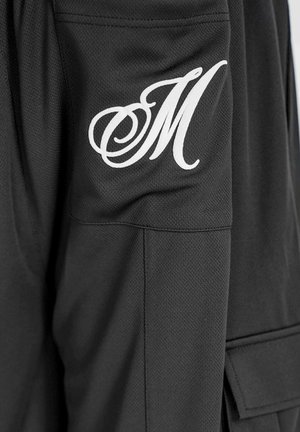Schwarze Sportjacke aus strukturiertem Stoff, mit einem großen weißen, geschwungenen "M" auf dem linken Ärmel und einer Pocket-Detail.
