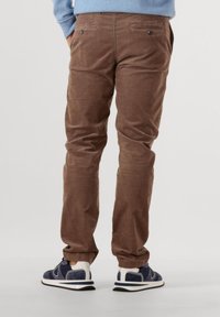 Man wearing brown corduroy trousers and navy blue and white sneakers, standing with hands in pockets, facing away from the camera.