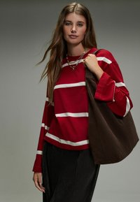 Rotes Langarmshirt mit weißen horizontalen Streifen, kombiniert mit einer dunklen Tasche. Das Model hält eine braune Tragetasche und trägt strukturierte schwarze Hosen. Minimale Accessoires.