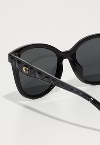 Lunettes de soleil noires avec des montures surdimensionnées et arrondies. Verres foncés et branches noires texturées présentent un accent logo subtil. Fini brillant tout au long.
