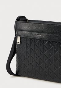 Svart lær kryssbody-veske med et teksturert diamantmønster og hevede sirkulære detaljer. Har en avtagbar stropp og merkevare-navn.