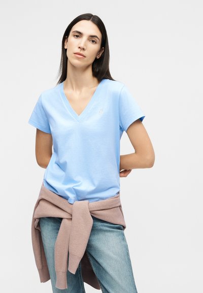 Γυναίκα με ίσια σκουρόχρωμα μαλλιά, που φοράει φανέλα με V λαιμόκοψη σε ανοιχτό μπλε χρώμα, ανοιχτό μπλε τζιν και μπεζ πουλόβερ δεμένο στη μέση της.