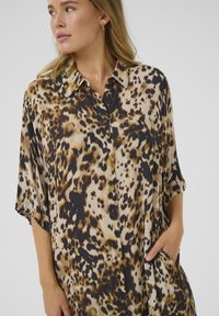 Camicia a maniche corte con stampa leopardata, colletto e tasche laterali, realizzata in tessuto morbido con diverse tonalità di marrone e nero.
