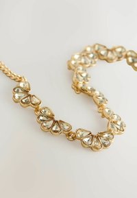 Goldene Halskette mit einem gemusterten Design, in das klare, tränenförmige Edelsteine in eine skulptierte Basis eingearbeitet sind. Sie zeigt ein glänzendes Finish und strukturierte Glieder.