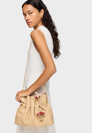 Mujer joven con cabello largo y oscuro lleva un vestido blanco sin mangas, sosteniendo un bolso de hombro beige suave con una flor decorativa y un charm dorado.