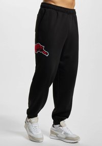 Svarta sweatpants med en broderad röd björnlogo på vänster lår, med dragsko i midjan och cuffade anklar, tillsammans med grå-och-vita sneakers.