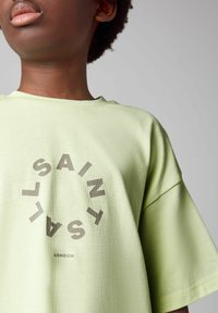 Svetlozelené oversized tričko vyrobené z bavlny, s centrálnym čiernym textovým potlačou, ktorý hovorí "SAINT LONDON".