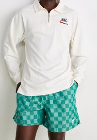 Vit långärmad zip-tröja med Nike-logotyp, tillsammans med gröna rutiga shorts med en vit Nike-swoosh. Mjukt material med avslappnad passform.
