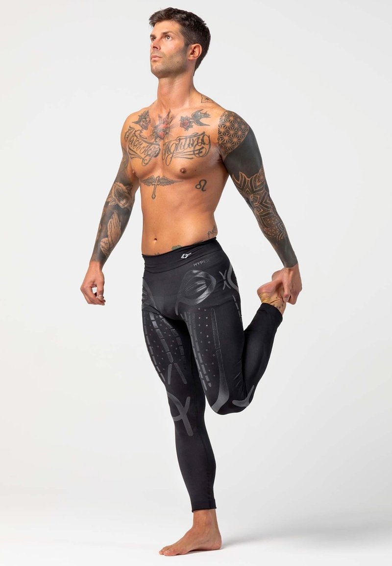 Leggings de sport noirs avec des motifs graphiques gris, dotés d'une taille haute. Le modèle étire une jambe tout en étant pieds nus.
