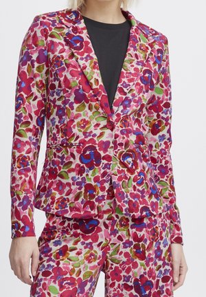 Woman wearing a colorful floral blazer and matching pants with red, purple, and green hues over a black top, standing against a white background.