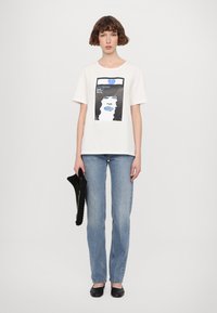 Weißes Baumwoll-T-Shirt mit Grafikdruck, der das Gesicht einer Frau zeigt, kombiniert mit hellblauen Jeans und schwarzen Ballerinas. Hält eine schwarze Tasche.