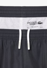 Melni brīvi pieguļoši sporta šorti ar baltu iekšējo jostasvietu. Iespējams, ar aukliņu aizdari un redzamu Lacoste logo uzkarinājumu. Audums ar sietu tekstūru.