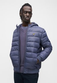 Navy blue puffer jacket with a zip front, ribbed cuffs, and a small gold logo. Worn over a purple sweater, with a textured finish.