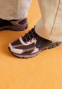 Smeđe i bež atletske cipele s mrežastim panelima, teksturiranom potplatom i crnim detaljima, uparene s svijetlim hlačama na žutoj površini.