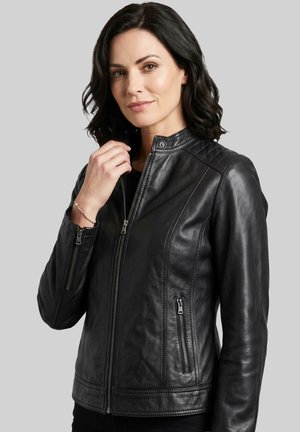 Woman with shoulder-length dark hair wearing a fitted black leather jacket with zippered pockets and a stand collar, posing against a gray background.