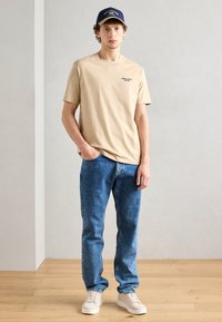 Beige bomulds T-shirt med et lille sort logo, parret med blå jeans. Iført en marineblå kasket og lyse beige sneakers på et trægulv.