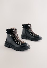 Black hiking boots with laces and metallic hardware, featuring a textured upper and fur lining, mounted on a thick, rugged sole.
