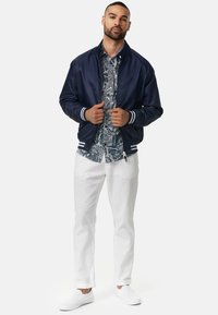 Giubbotto bomber navy con polsini a strisce bianche, camicia button-up a fantasia, pantaloni bianchi e scarpe slip-on. Elementi di abbigliamento casual combinati.