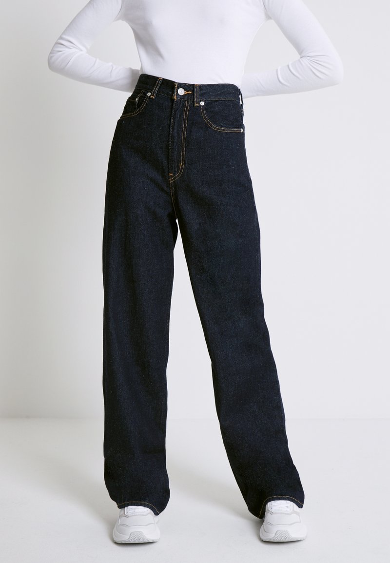Personne portant un jean large taille haute bleu foncé, un haut blanc à manches longues et des baskets blanches, debout les mains sur les hanches.