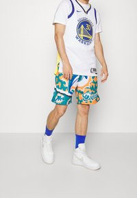 Persona che indossa una maglia dei Golden State Warriors, pantaloni corti colorati dei Miami Dolphins, calzini blu e scarpe Nike bianche, in piedi su uno sfondo neutro.
