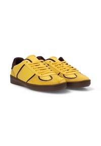 Las zapatillas de ante amarillas cuentan con una puntera de cuero suave, acentos en marrón y una suela de goma marrón. Tienen cordones tradicionales y detalles perforados.
