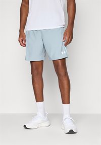 Vīrietis, kurš valkā gaiši zilas Under Armour sporta šortus, baltas sporta zeķes, baltus sporta apavus un baltu krekli. Viņš stāv uz viendabīga fona.