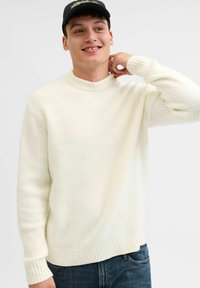 Suéter blanco de punto acanalado con un cuello alto y mangas largas, combinado con jeans oscuros; modelo con una gorra negra.
