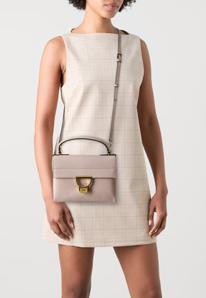 Borsa in pelle rosa con design strutturato, accessori in oro e tracolla rimovibile, tenuta da una persona che indossa un vestito beige a fantasia.