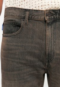 Fekete farmernadrág, mely kopott hatású, gombos záródással és egy kis kék címkével rendelkezik. Klasszikus öt zsebes dizájn, enyhén fakult felülettel.