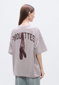 Gray oversized t-shirt featuring "PIROUTTES" text and graphic of ballet shoes on the back. Soft cotton material. Casual design with dropped shoulders.