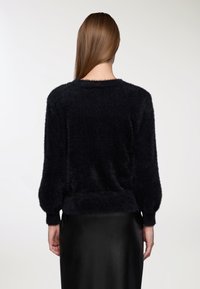 Kobieta z prostymi brązowymi włosami, ubrana w czarny puszysty sweter z długim rękawem i czarną skórzaną spódnicę, pokazana od tyłu.