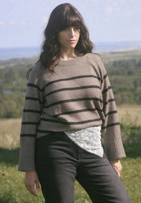 Pull en stripe grise avec des manches larges, associé à un jean foncé avec un accent brodé blanc au niveau de la taille. Cadre naturel en extérieur.