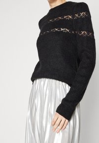 Maglione nero a maglia con dettagli in pizzo aperto sulle spalle, scollo rotondo e maniche lunghe, abbinato a una gonna plissettata argento metallica.