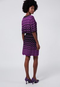 Abito a zigzag viola e nero con maniche corte, vita aderente e gonna svasata, abbinato a scarpe col tacco viola.