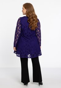 Paarse kanten tuniek met bloemenpatroon, lange mouwen en een uitlopend zoom. Gemaakt in combinatie met zwarte broek en zwarte hakken.