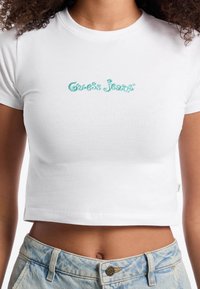 Bijela pamučna kratka majica s turkiznim izvezenim logotipom "Guess Jeans" na prednjoj strani. Ima klasičan okrugli izrez i kratke rukave.