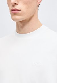 Κοντινό πλάνο ενός ατόμου που φοράει μια απλή λευκή μπλούζα με στρογγυλή λαιμόκοψη και ένα διακριτικό κέντημα με το λογότυπο "BOSS" στο στήθος.