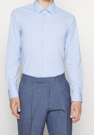 Mężczyzna w jasnoniebieskiej, długorękawowej koszuli z guzikami, wpuszczonej w niebieskie teksturowane spodnie wizytowe, stoi z rękami swobodnie opuszczonymi wzdłuż ciała.