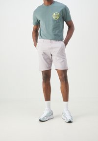 Shorts gris clair avec des revers, associés à un t-shirt vert avec un imprimé graphique jaune sur la poitrine, et des chaussures de sport blanches.