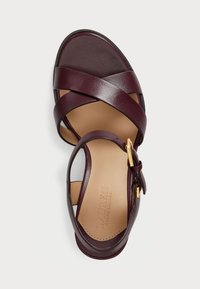 Burgundi színű bőr szandál keresztpántos dizájnnal és arany csattal. Simább felsőrész lapos talppal, kontrasztos barna talpvéggel.