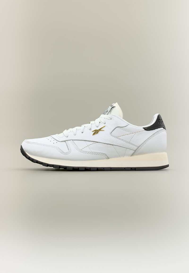 Λευκό χαμηλό αθλητικό παπούτσι Reebok με μαύρη ταμπέλα στη φτέρνα, χρυσή διακόσμηση λογότυπου, λευκά κορδόνια και μαύρη ελαστική σόλα σε ουδέτερο φόντο.