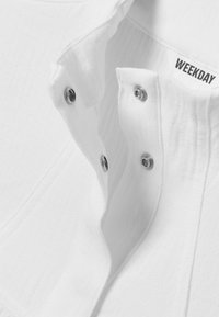 Chemise blanche en coton avec un col à revers, dotée de boutons-pression argentés et d'une étiquette portant la mention « WEEKDAY » près du col.
