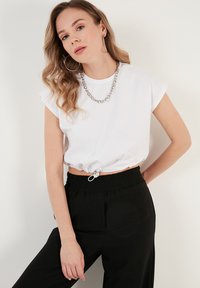 Weißes, kurzes T-Shirt mit kurzen, umgeschlagenen Ärmeln, gerafftem Saum mit einem Zugband, kombiniert mit schwarzen, elastischen Hosen. Silberne Kettenhalskette.