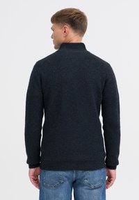 Dunkelblauer Strickpullover mit hohem Kragen, strukturiertem Stoff und langen Ärmeln. Getragen mit hellblauen Jeans, Sicht von hinten.