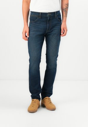 Jeans de denim azul oscuro con un ajuste clásico, diseño de cinco bolsillos y detalles de costura sutiles, combinados con zapatillas color crema.