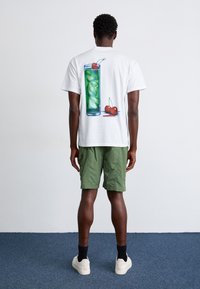 Balti trumpomis rankovėmis marškinėliai su spalvingu aukšto stiklinio gėrimo su ledu ir vyšniomis atvaizdu nugaroje, derinami su žaliais šortais.