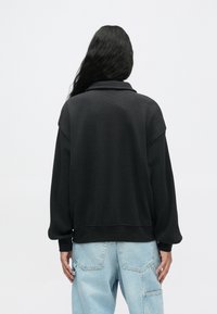 Pessoa com cabelo longo e escuro vestindo uma blusa preta texturizada de mangas compridas e jeans azul claro, de costas contra um fundo simples.